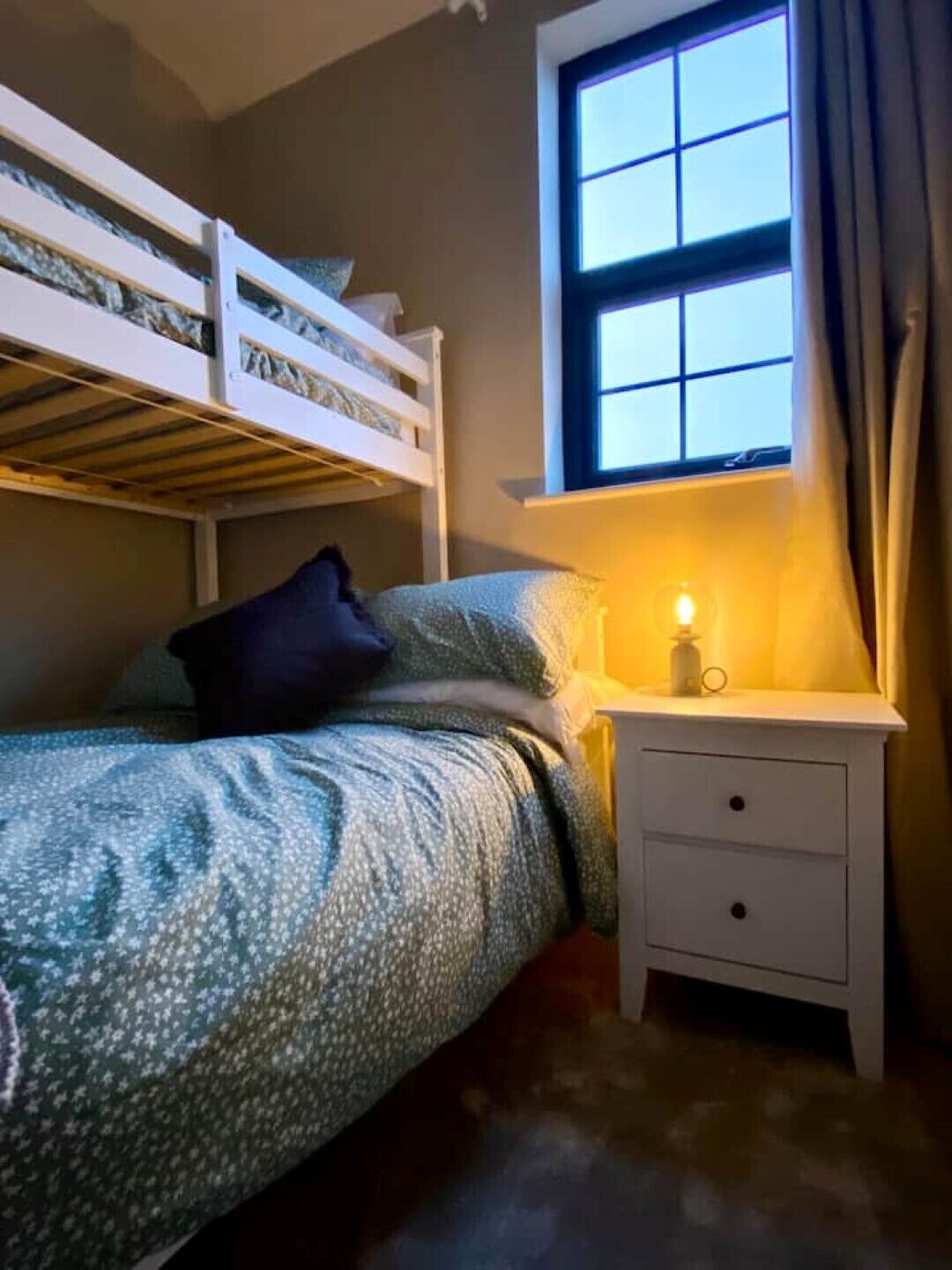 Una camera da letto con un letto a castello, un comodino con una lampada e una finestra con tende.