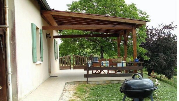Une terrasse extérieure couverte, équipée d’un barbecue, de meubles en bois et offrant une vue sur une maison aux volets verts.