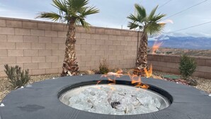 Una chimenea exterior circular con leña ardiendo, rodeada por un muro de ladrillo y palmeras.