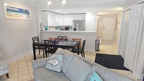 Uma sala de estar com um sofá cinzento, uma mesa de jantar com cadeiras, uma área de cozinha com armários brancos e uma porta que dá para outro quarto.