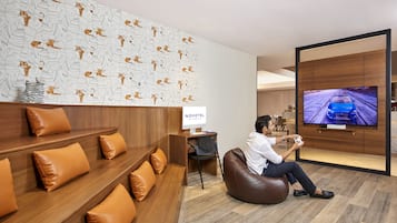 A modern hotel lobby with a wooden reception desk, a Novotel sign, a large TV screen, and a seating area with leather cushions.
