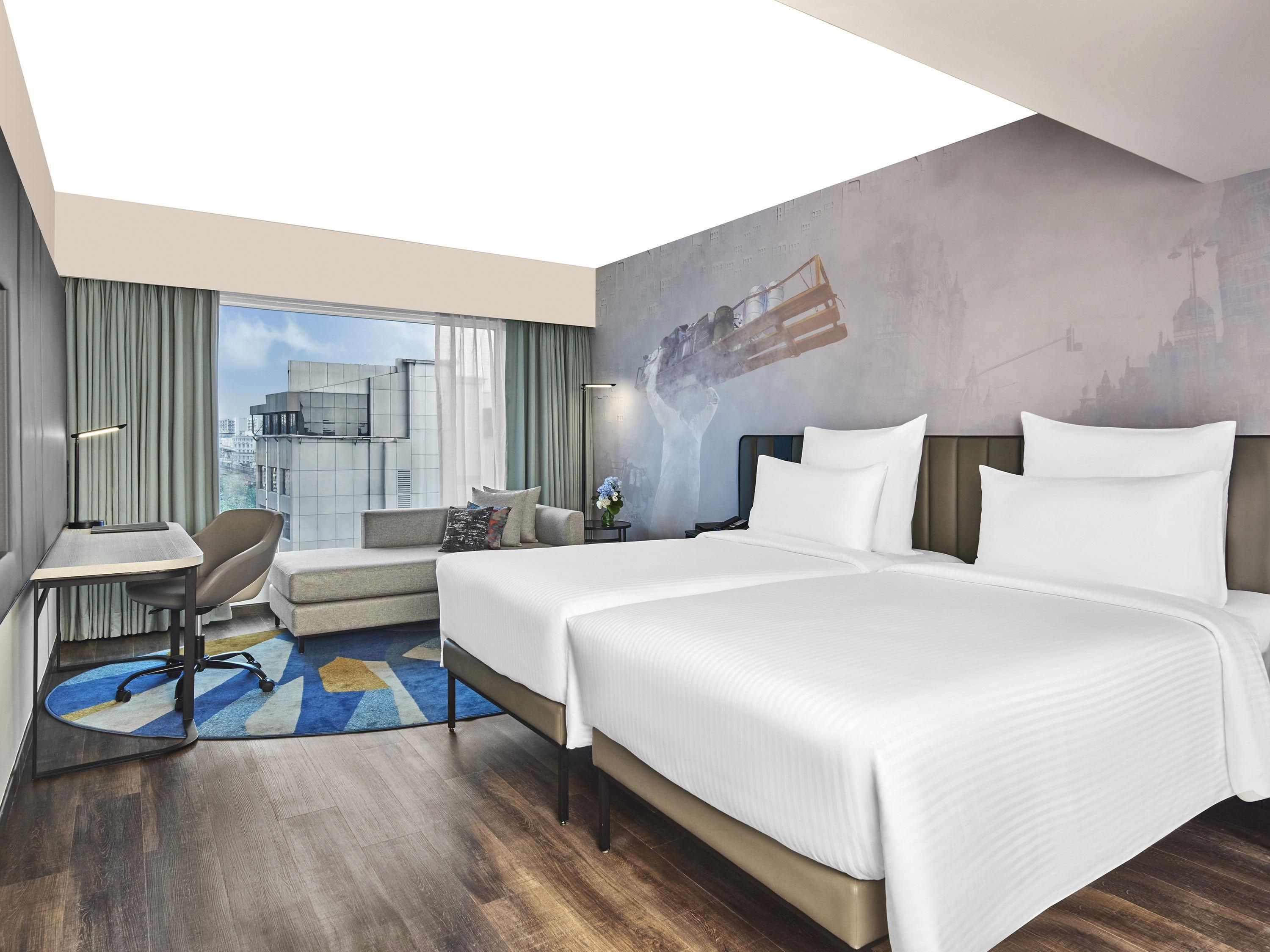A modern hotel room with two beds, a desk, a chair, and a large window offering a city view.