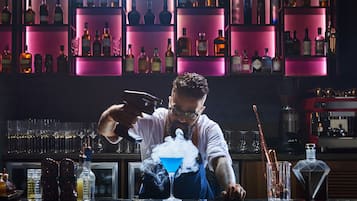 Un bar con estantes llenos de botellas y vasos, un barman preparando una bebida, y un coctelera sobre la barra.