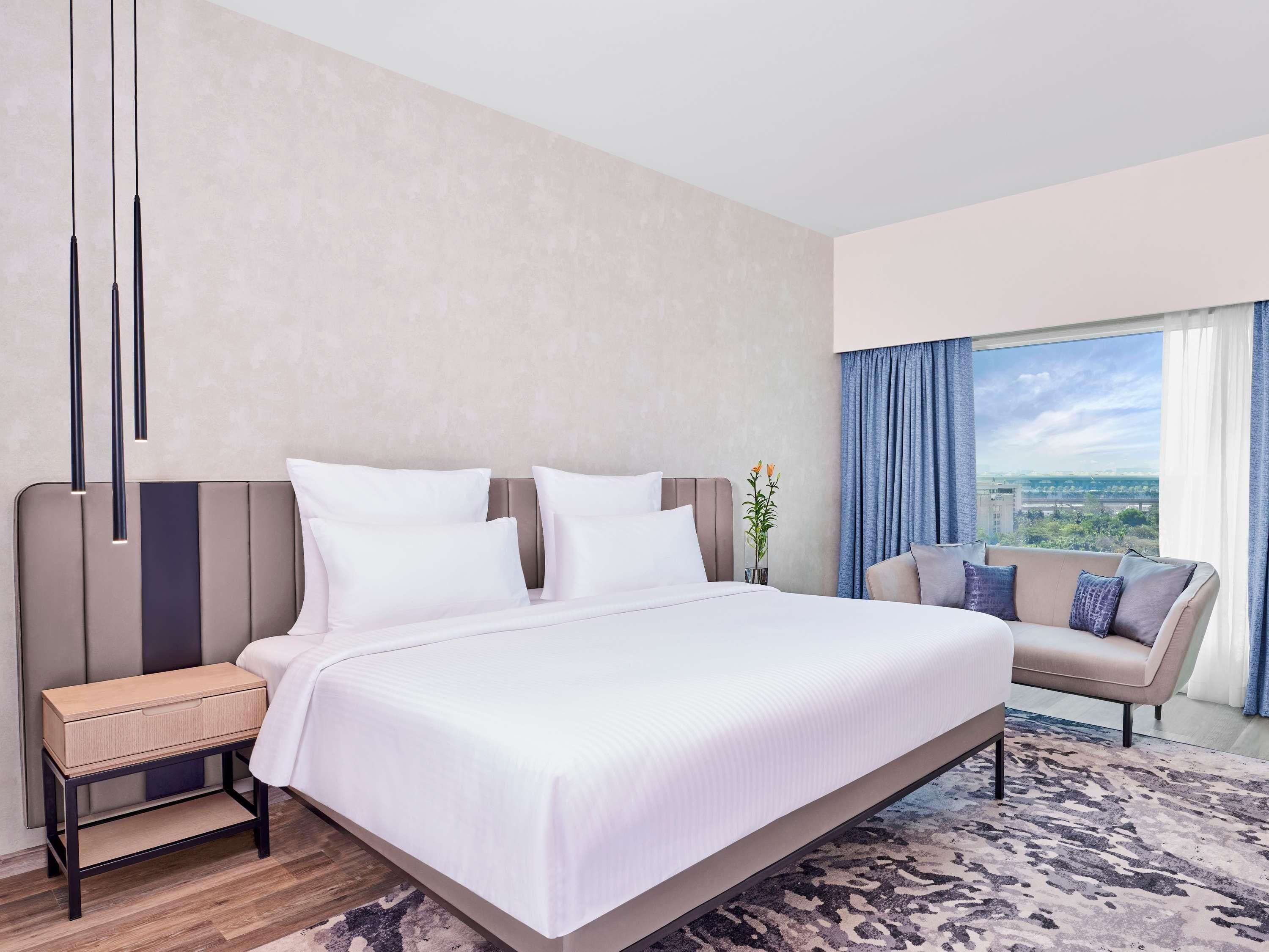 A modern hotel room with a large bed, a nightstand, a sofa, a view of the city through the window, and a vase with flowers.