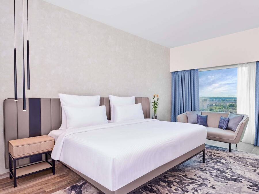 Habitación de hotel moderna con una cama grande, una mesita de noche, un sofá, vistas de la ciudad a través de la ventana y un florero con flores.