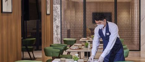 A restaurant with green upholstered chairs, neatly set dining tables, and a waiter attending to a table.