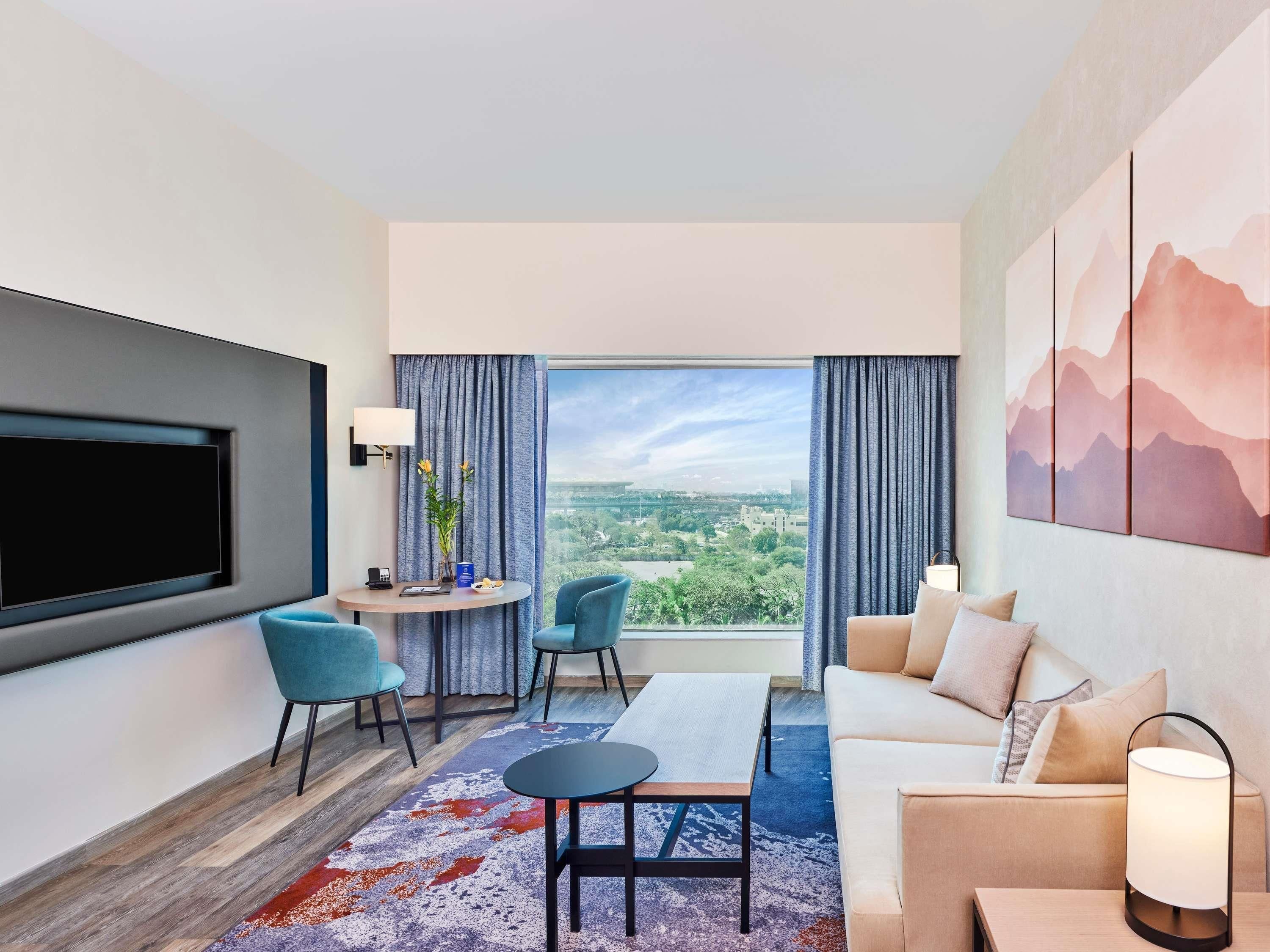 A modern hotel room with a large TV, a sofa, a dining table, and a view of the cityscape.