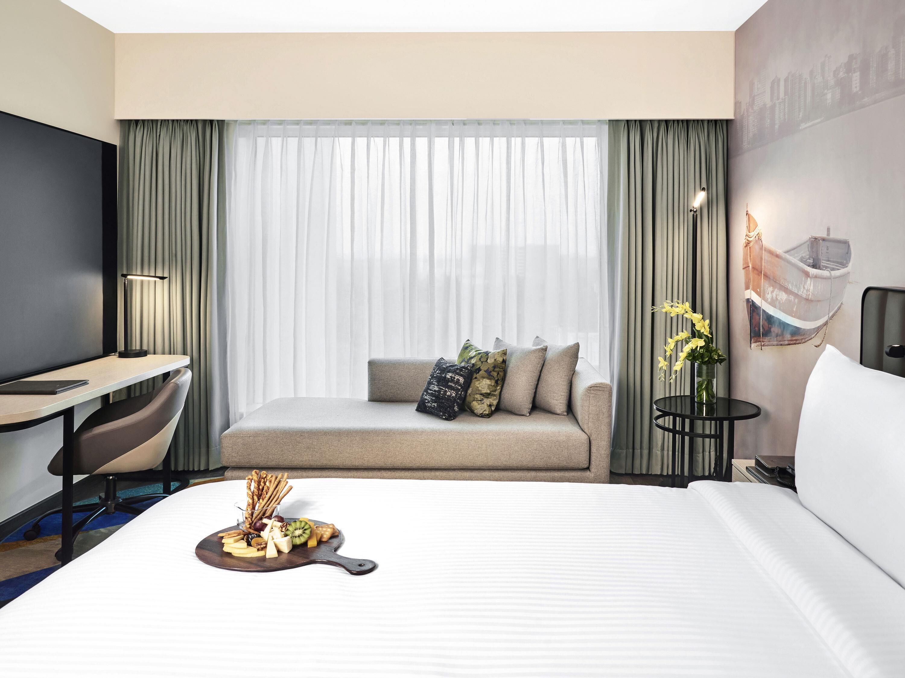 A hotel room with a bed, a desk, a chair, a small table with flowers, and a wall-mounted TV.