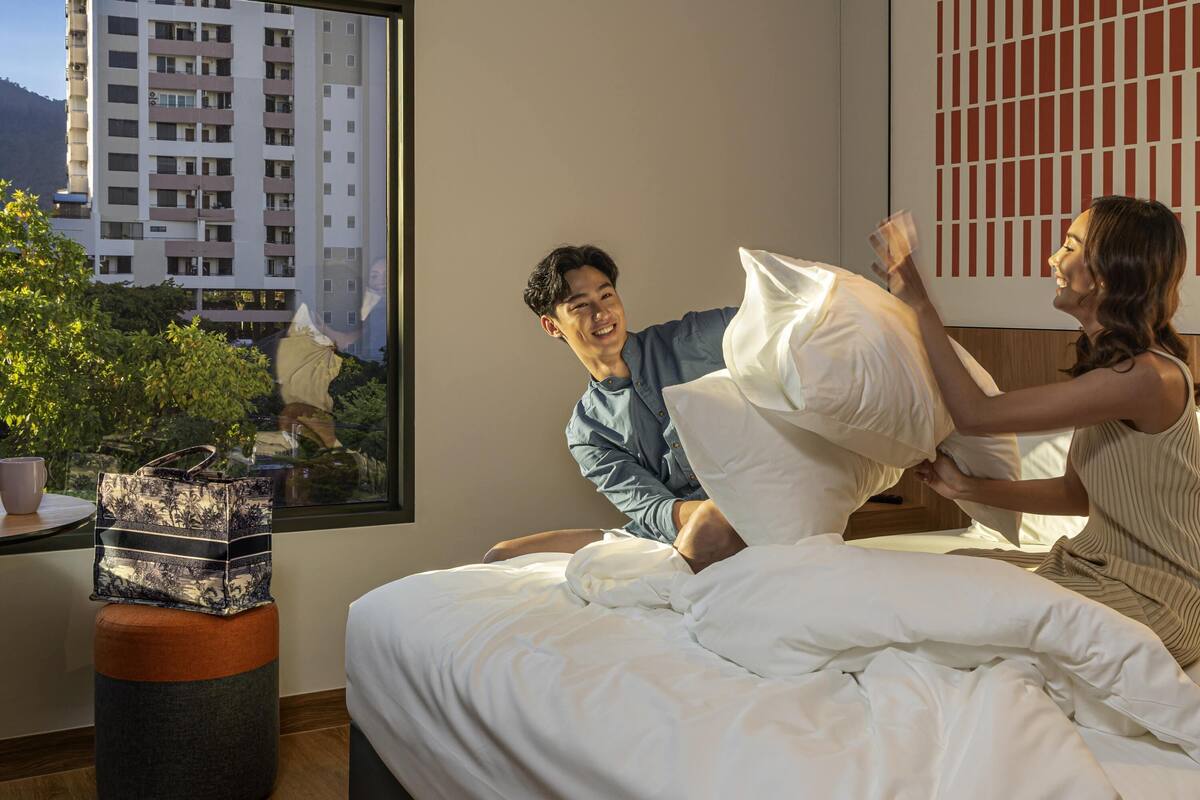Un dormitorio con cama, ventana con vista a edificios y una mesita con planta y bolso.