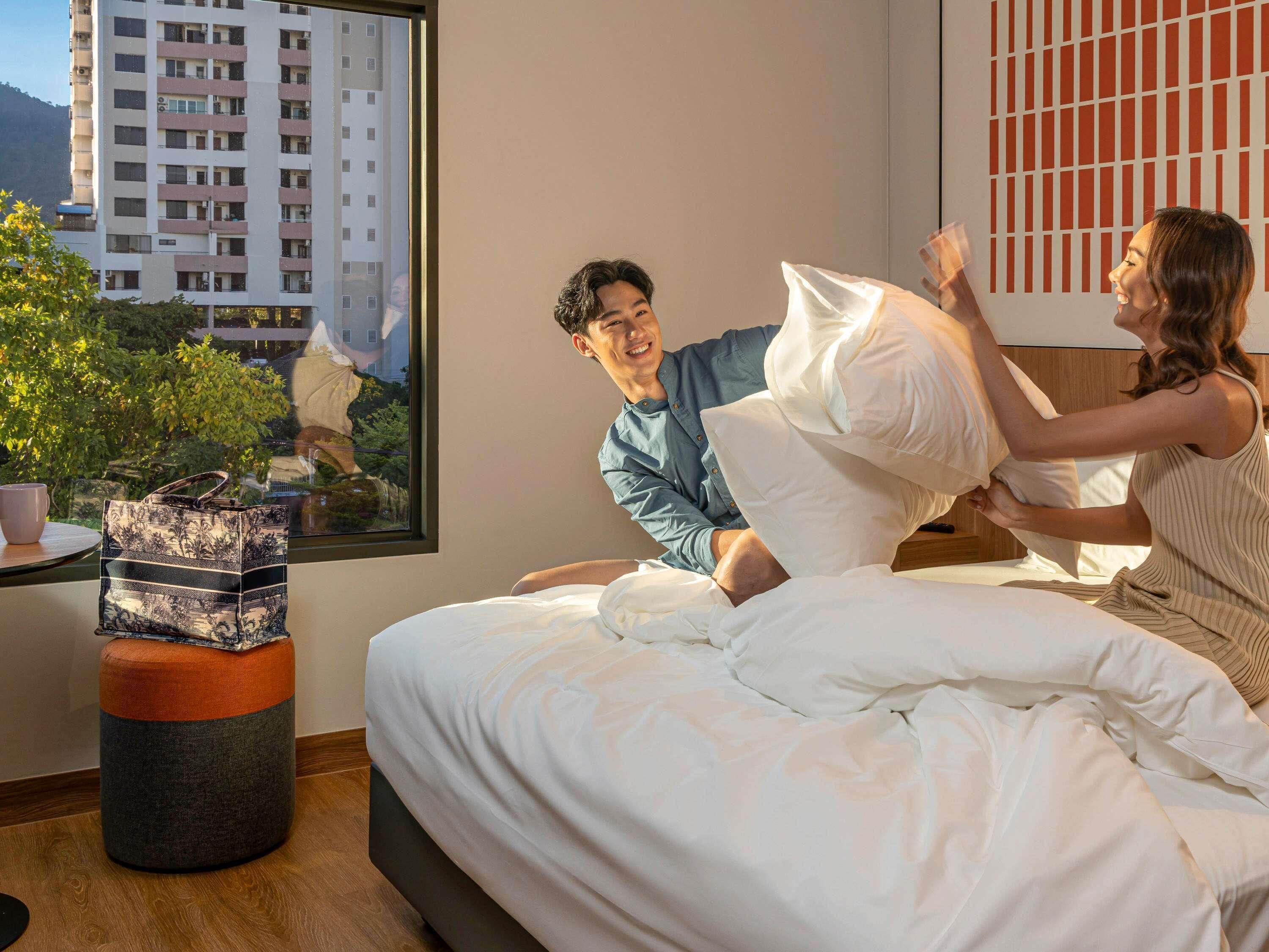 Una habitació amb un llit, una finestra amb vista a edificis, una taula petita amb una tassa i un sac decoratiu.