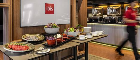A buffet setup with various food items and a large screen displaying the ibis logo.