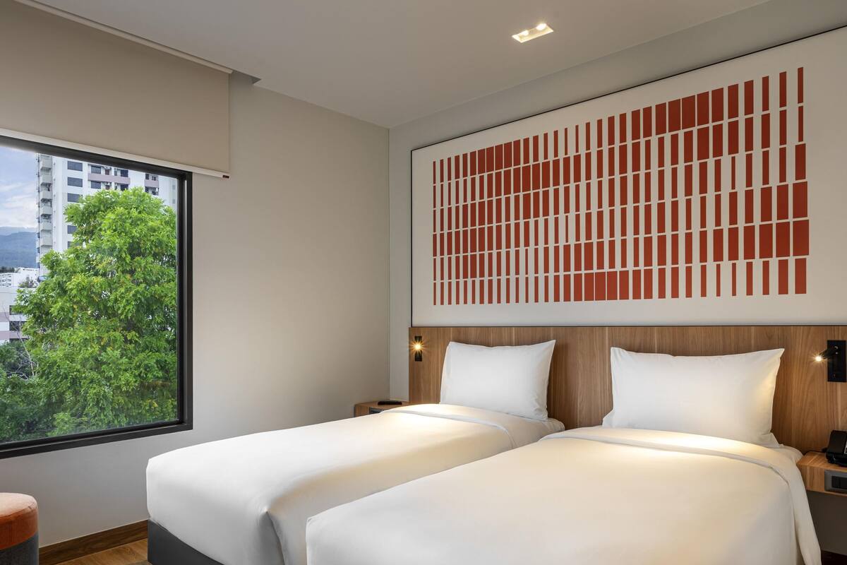 Habitación de hotel con dos camas, un ventanal con vistas a edificios y árboles, y un panel decorativo en la pared.
