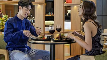 Um homem e uma mulher jantando à mesa, com prato, copos de vinho e uma pequena planta.