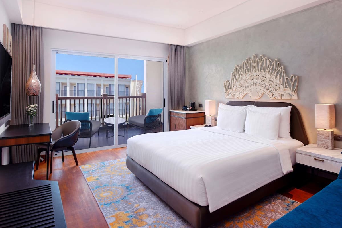A hotel room with a large bed, a desk with a chair, a TV, a balcony with a view of buildings, and a decorative wall piece.