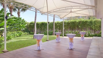 An outdoor covered area with tables and chairs, surrounded by greenery and a clear sky.