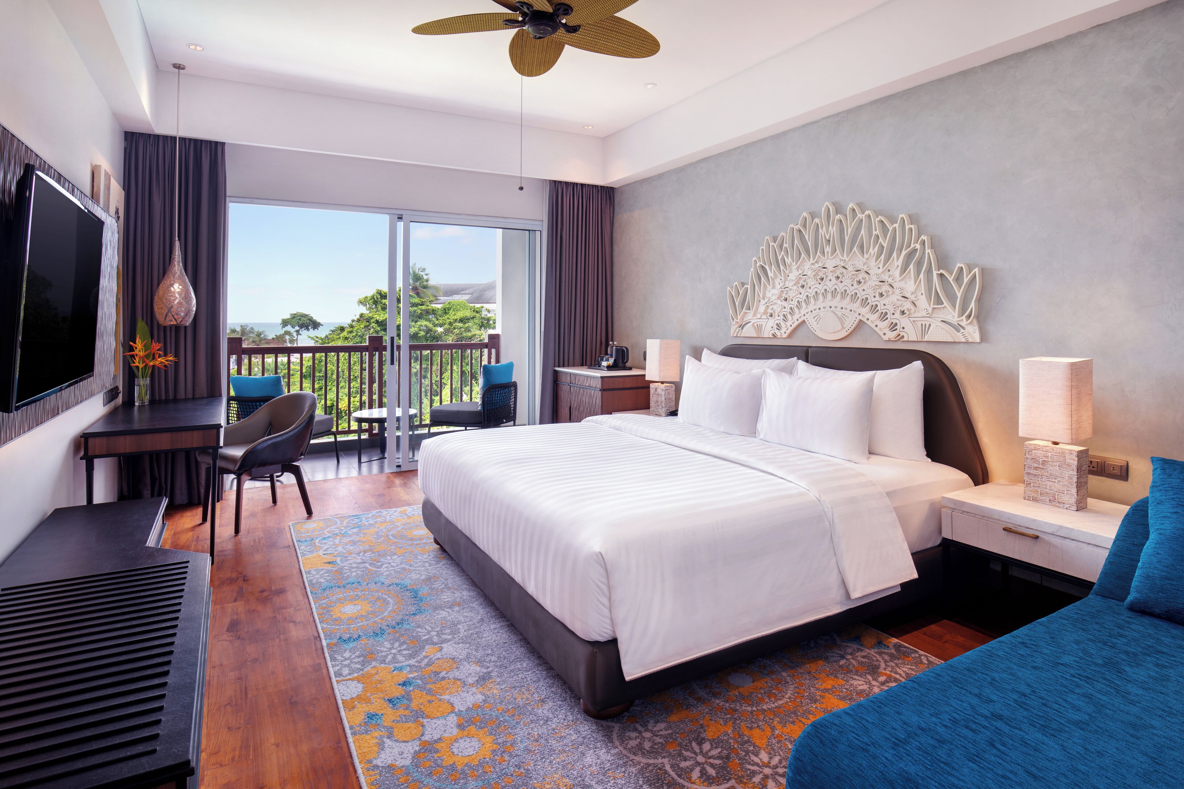 A modern hotel room with a large bed, a desk, a chair, a TV, and a balcony with a view.