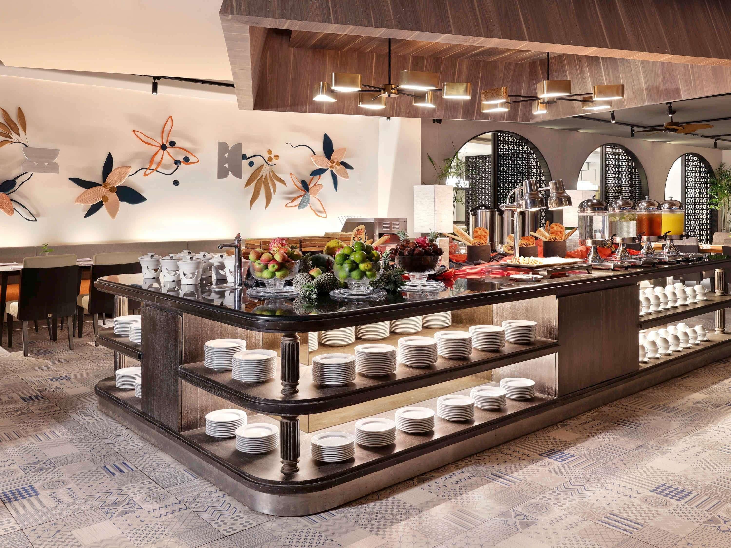 A buffet area with a variety of food items, including fruits and beverages, and a counter with stacked plates.