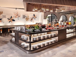 Uma área de buffet com diversos tipos de comida, incluindo frutas e bebidas, e um balcão com pilhas de pratos.