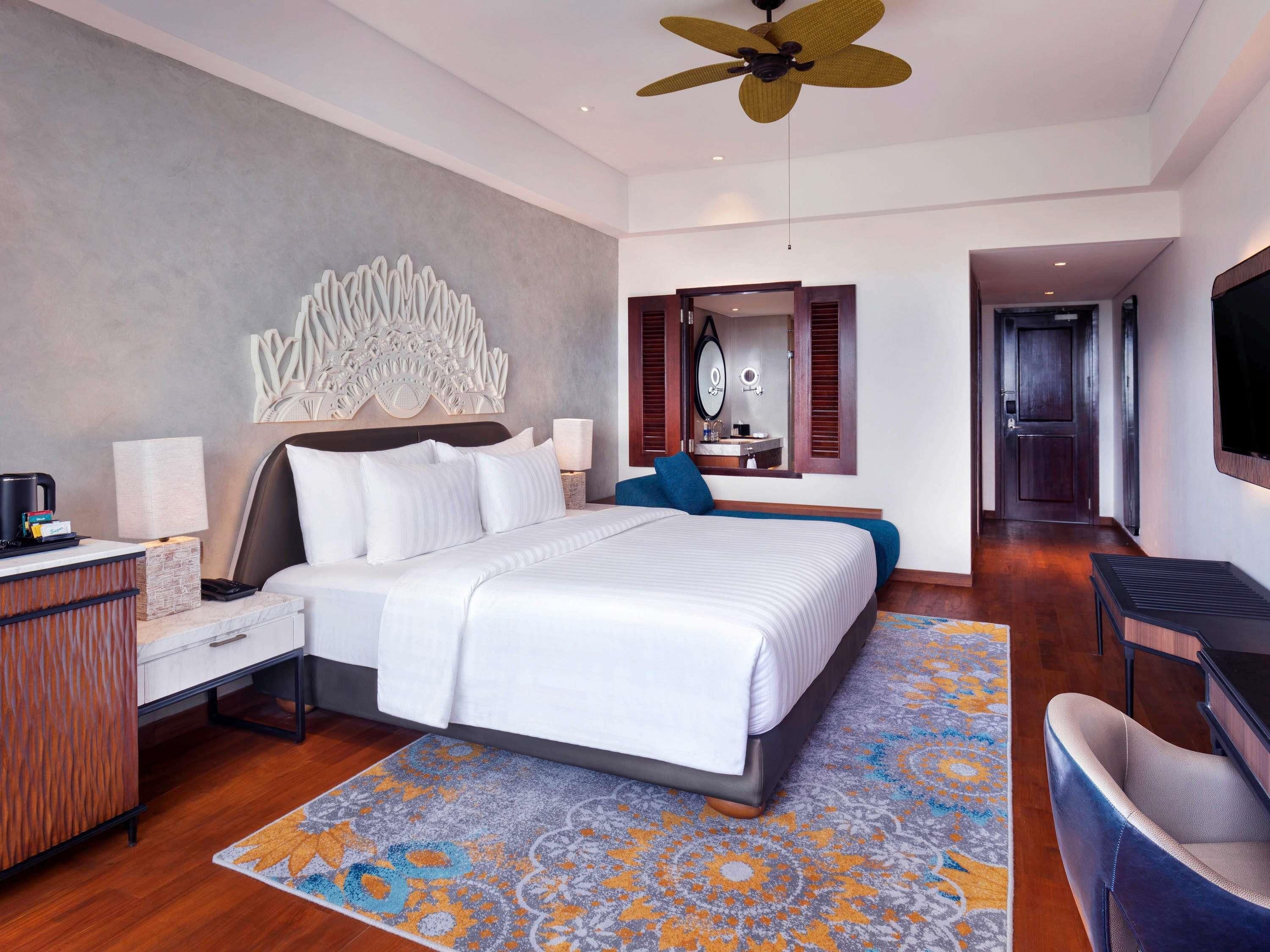 A modern hotel room with a large bed, a desk with a lamp, a ceiling fan, and a large mirror.