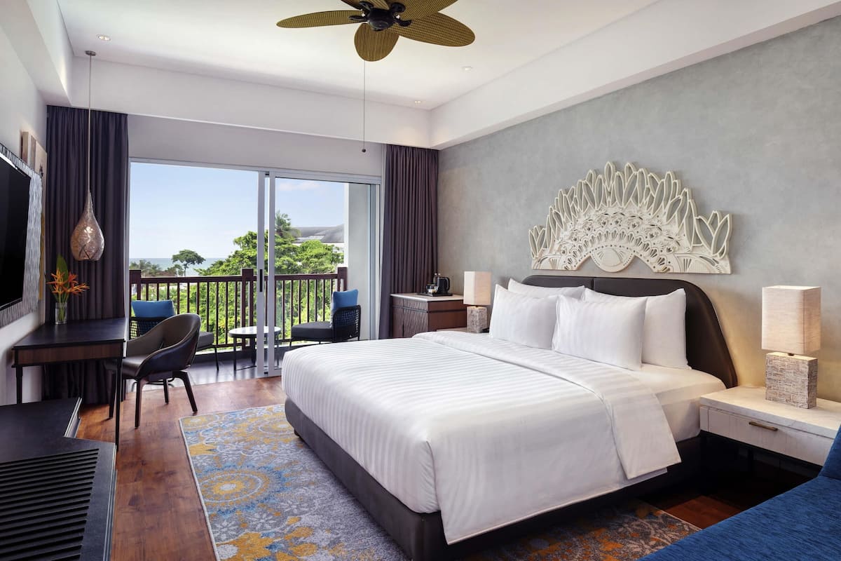 A hotel room with a large bed, a desk, a chair, a TV, a balcony with a view, and a decorative wall piece.