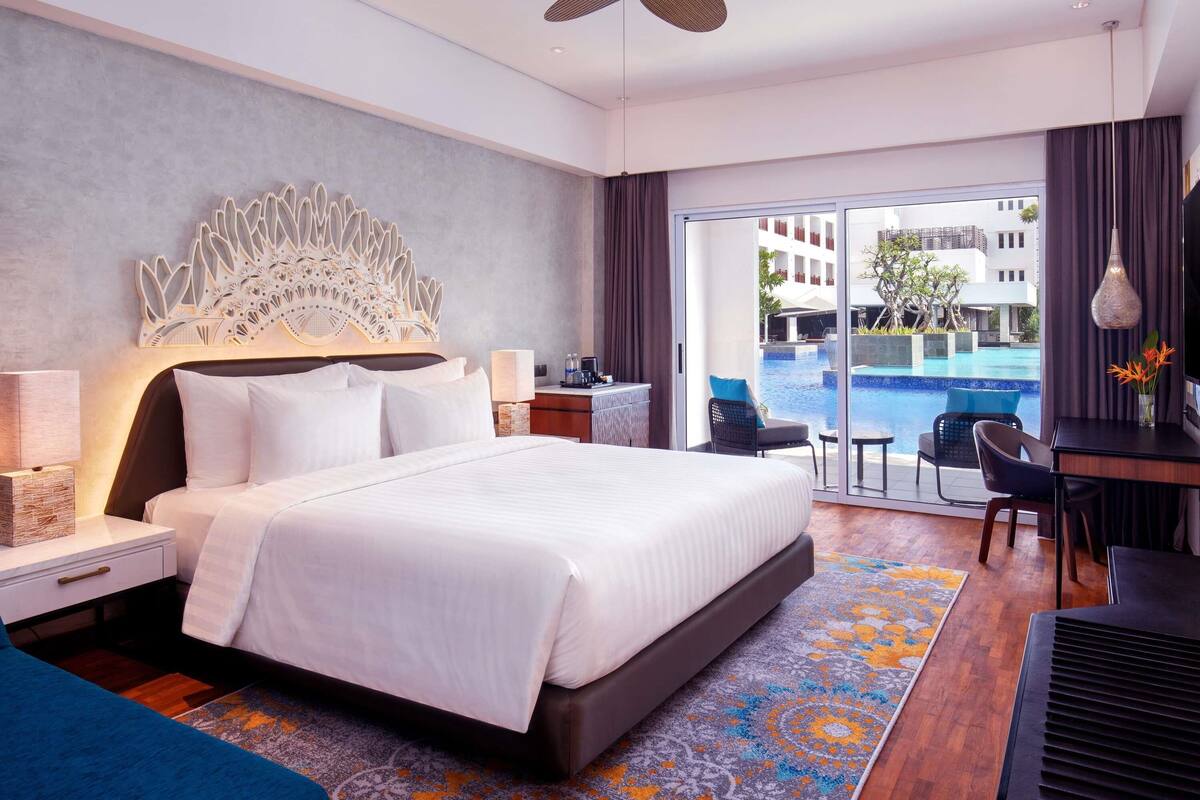 A hotel room with a bed, a desk, a chair, a nightstand, a lamp, a ceiling fan, and a view of a swimming pool.