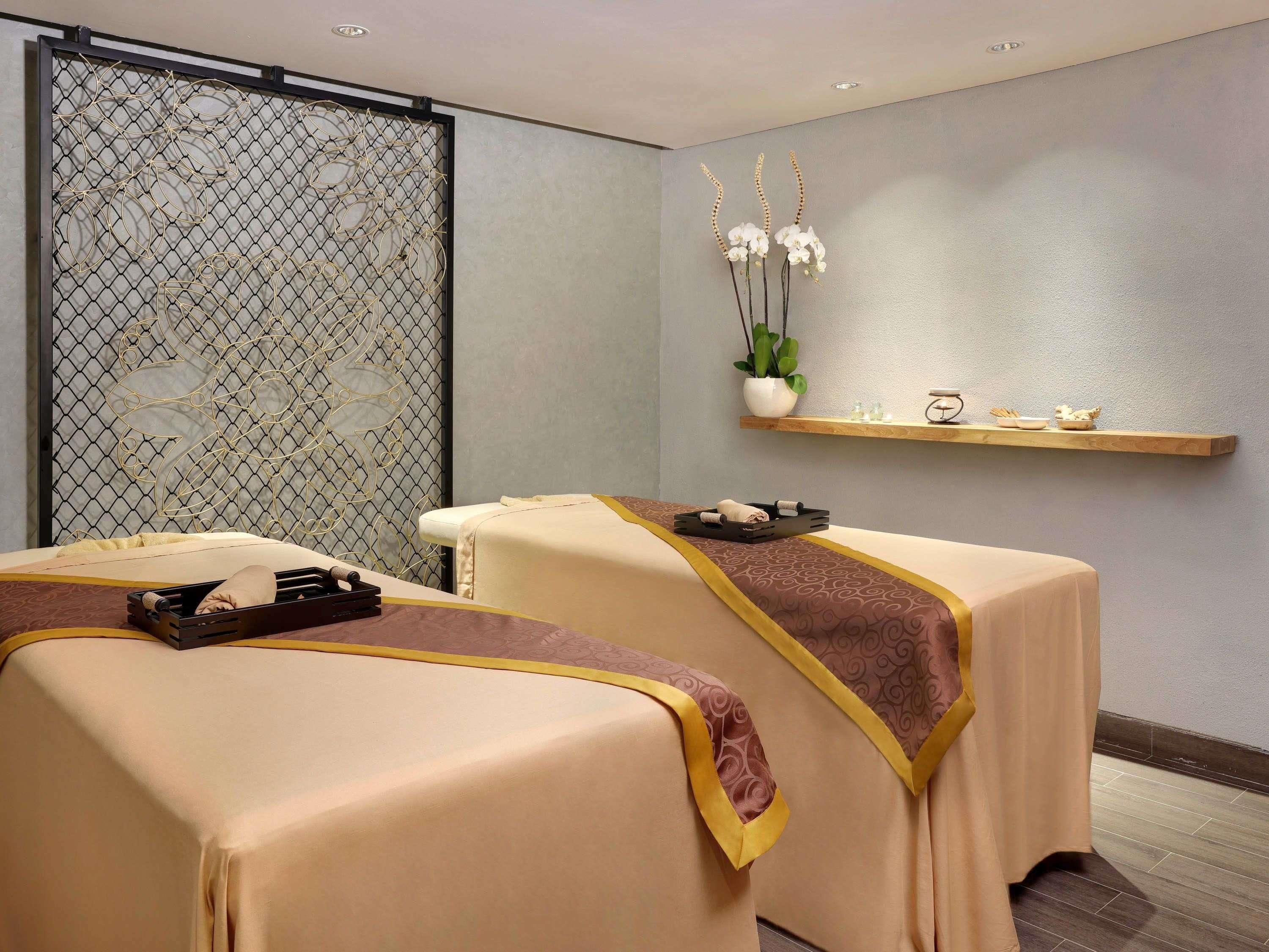 Un cuarto de spa con dos camillas de masaje, un muro decorativo, una estantería con artículos de tocador y una planta en maceta.