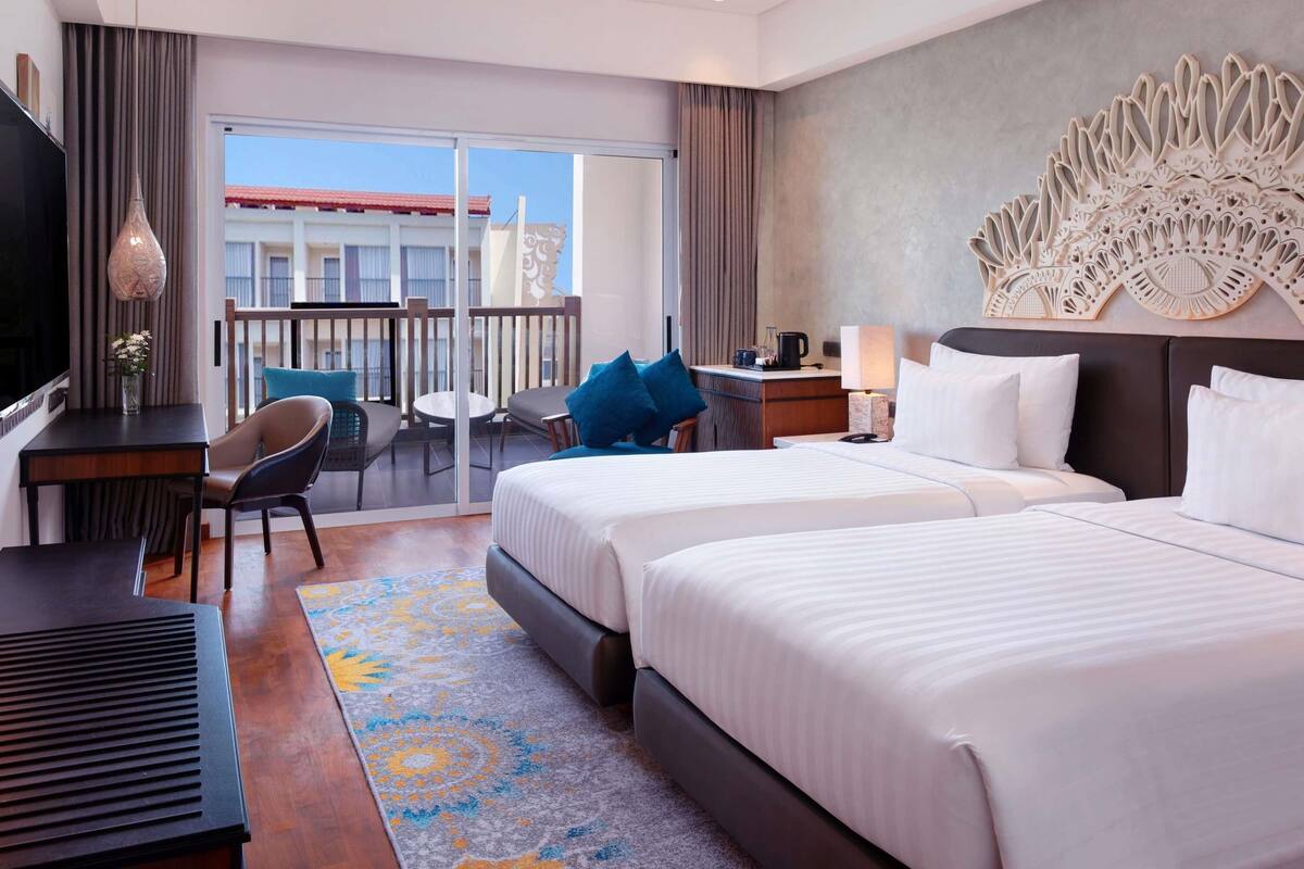 A hotel room with two beds, a desk, a chair, a TV, and a balcony with a view.