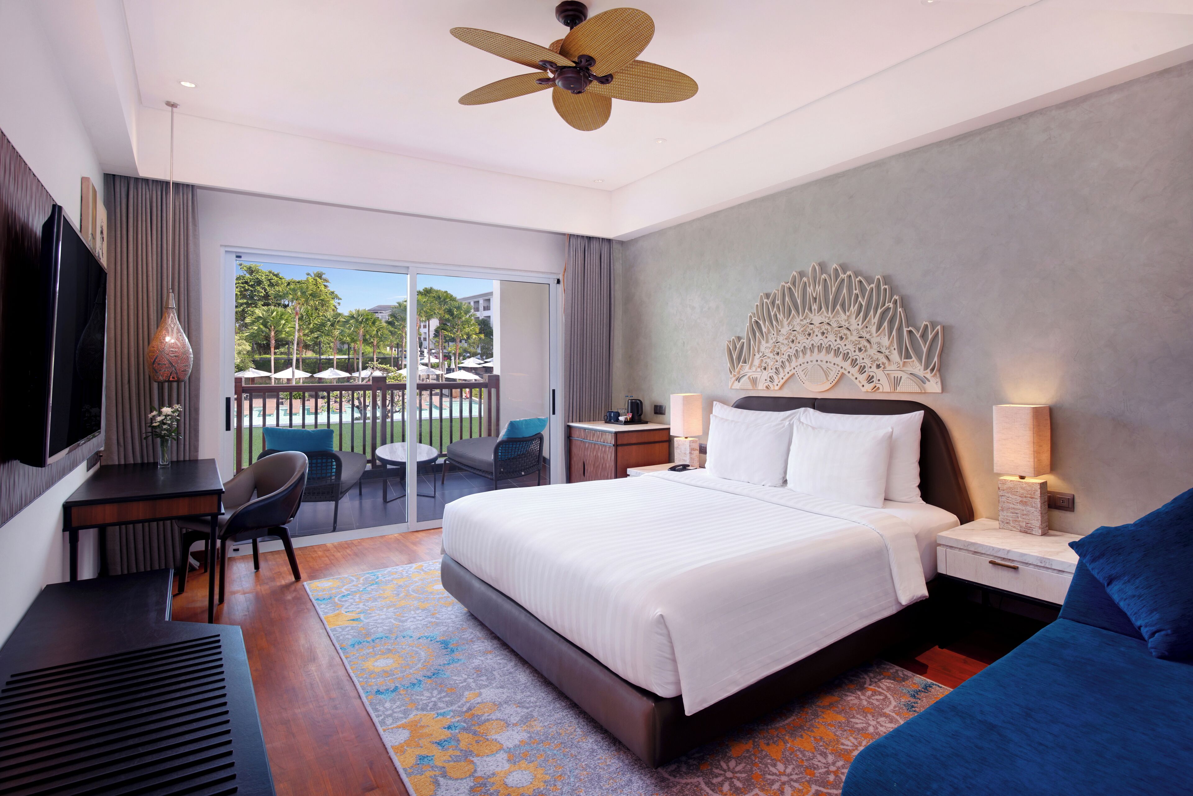 Una habitación de hotel moderna con una cama grande, un escritorio, una silla, un balcón con vistas a palmeras y un ventilador de techo.