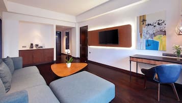 A modern living room with a grey sofa, a wooden coffee table, and a blue chair. There is a flat-screen TV mounted on the wall and a painting above it.