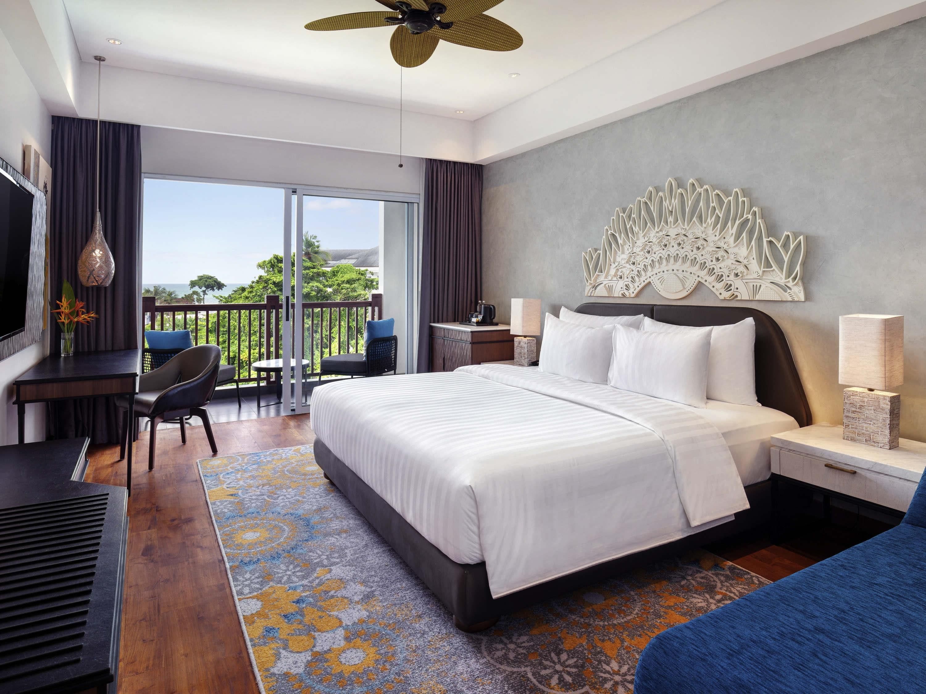 A hotel room with a large bed, a desk, a chair, a TV, a balcony with a view, and a decorative wall piece.