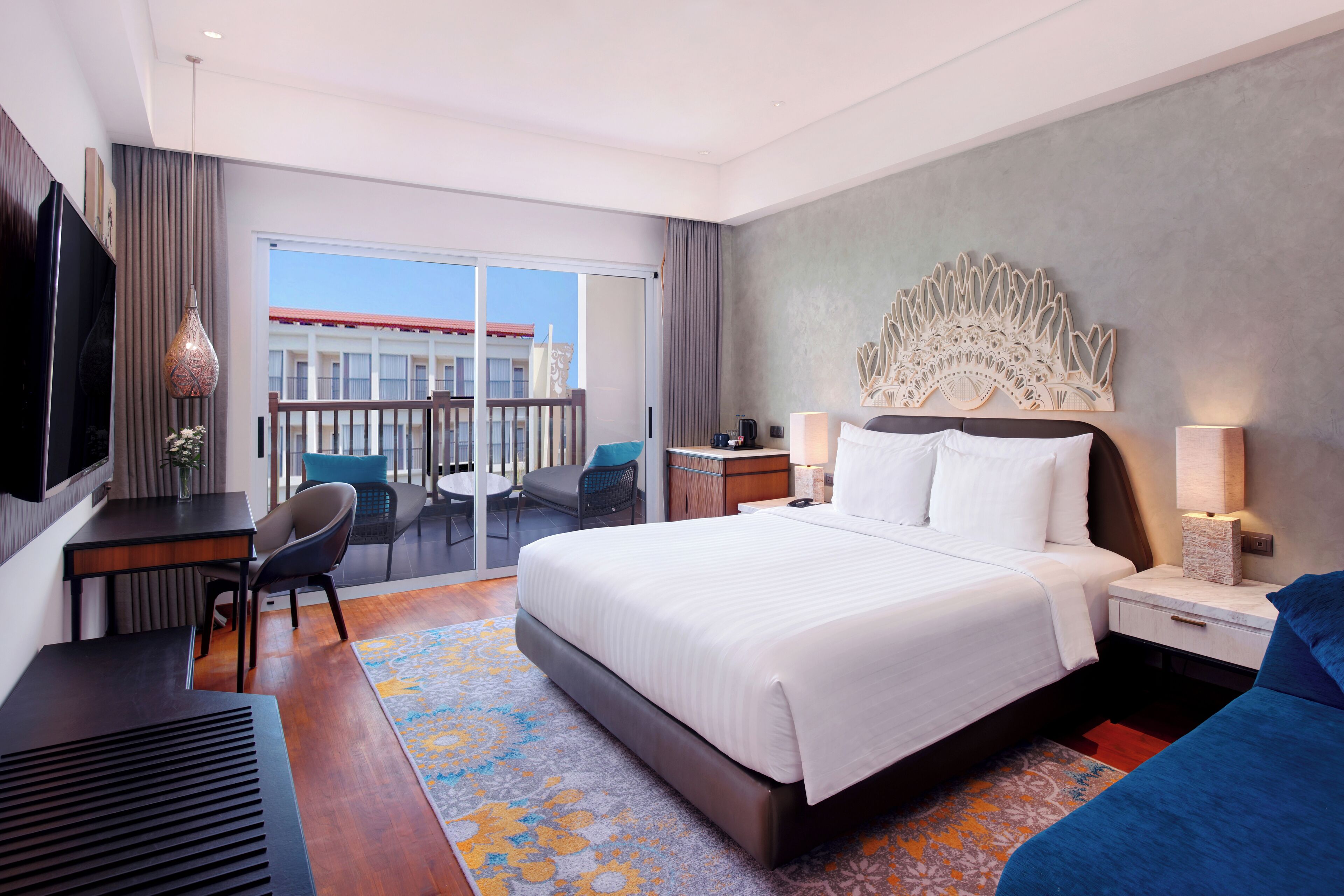 A hotel room with a large bed, a desk, a chair, a TV, and a balcony with a view of a building.