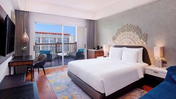 A hotel room with a large bed, a desk, a chair, a TV, and a balcony with a view of a building.