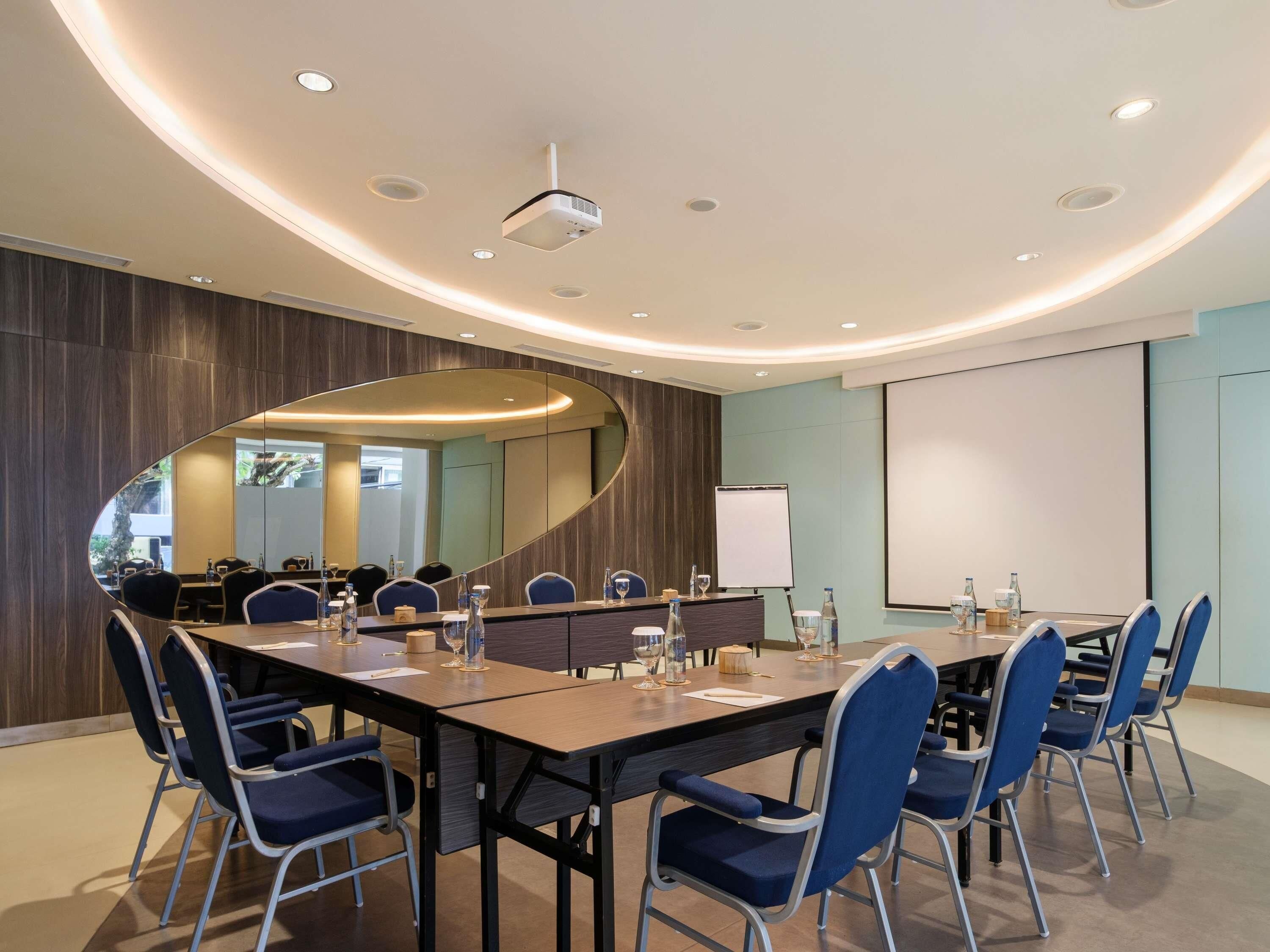 Ein Konferenzraum mit langem Tisch, blauen Stühlen, einem Projektor und einer Tafel.