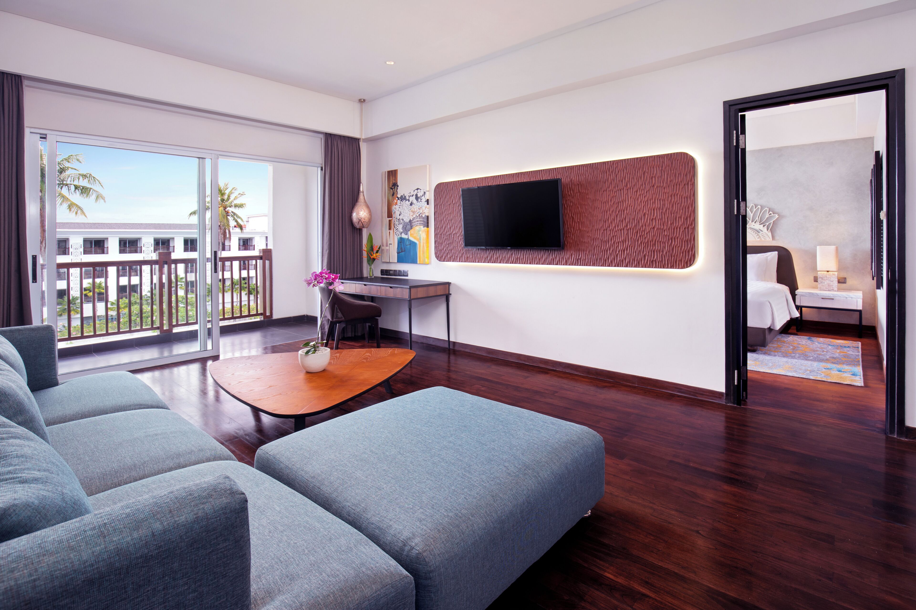 Una habitación de hotel moderna con un sofá grande, una mesita con un florero, un televisor de pantalla plana en una pared texturizada y un balcón con vistas a palmeras.
