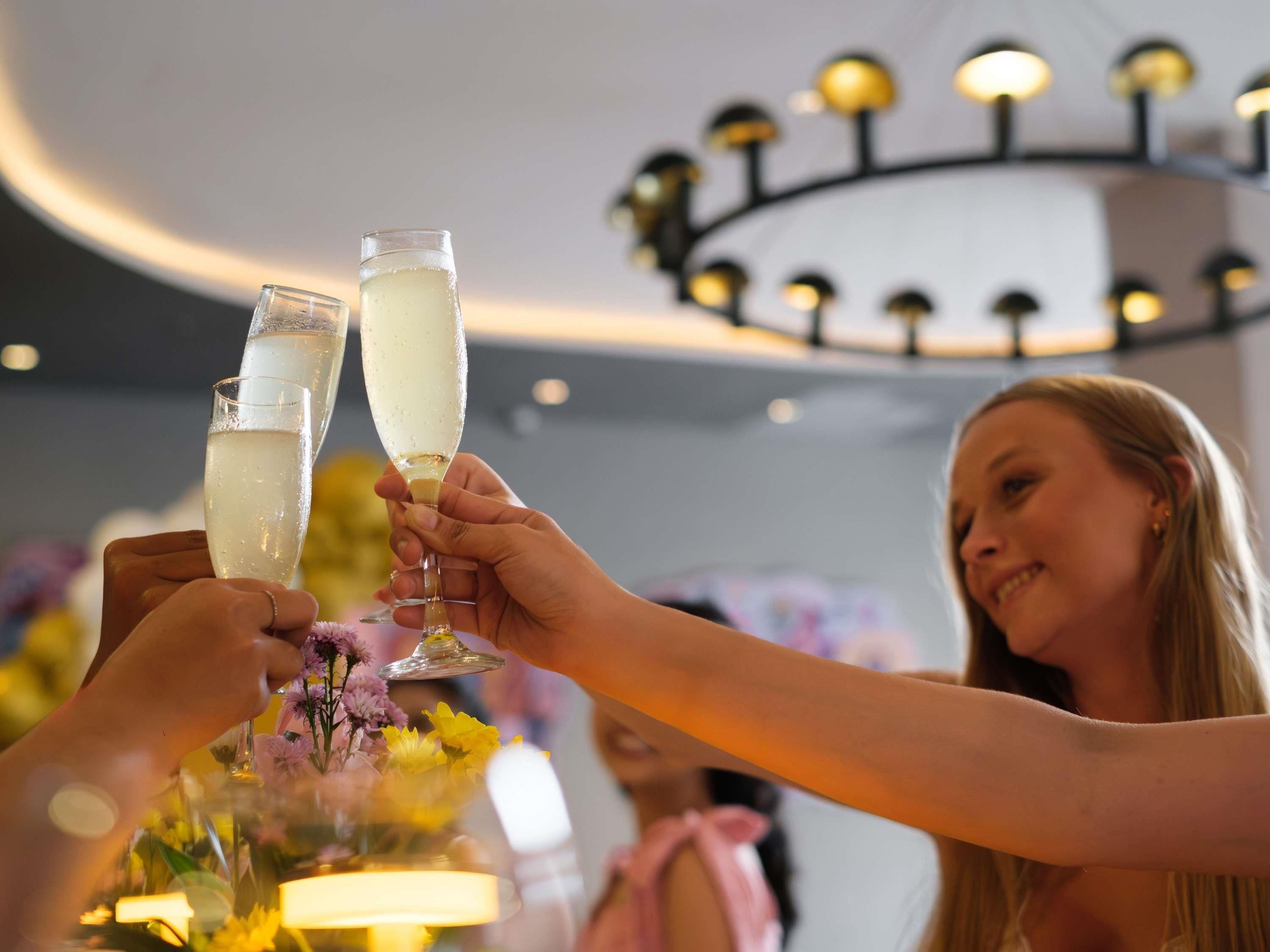 Twee handen vasthouden champagneglazen, een kroonluchter en een tafel met bloemen.