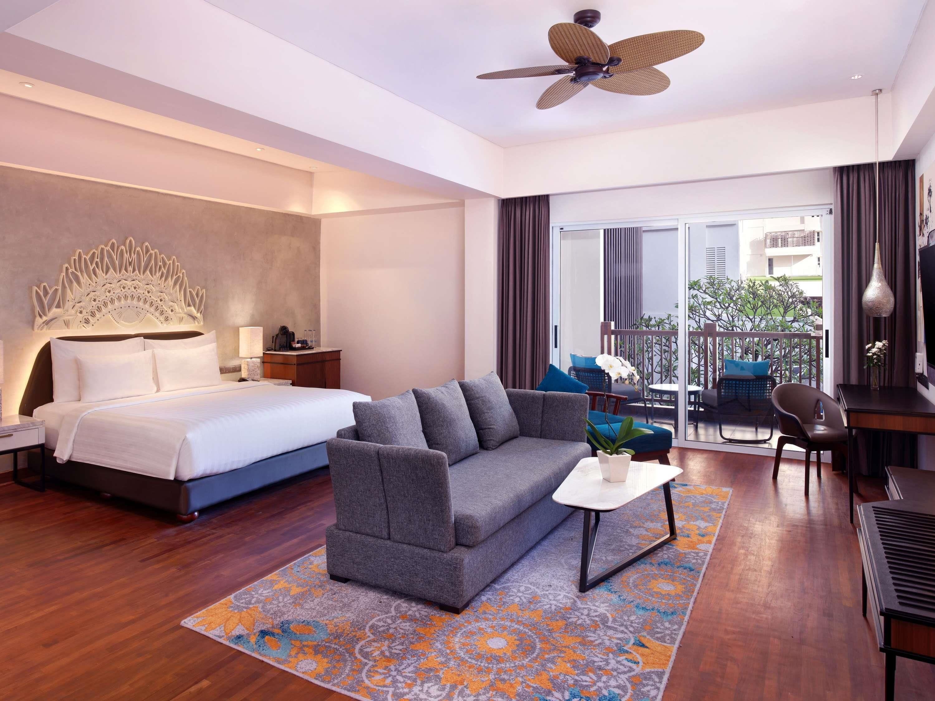 A modern hotel room with a large bed, a grey sofa, a small table, a ceiling fan, and a balcony with outdoor seating.
