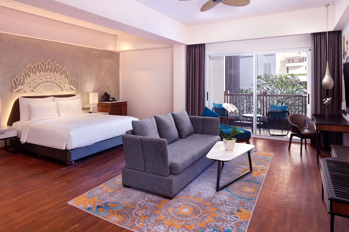 Quarto de hotel moderno com uma cama grande, um sofá cinzento, uma mesa pequena, um ventilador de teto e uma varanda com área de estar ao ar livre.