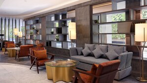 A modern lobby with a grey sofa, brown leather chairs, a glass coffee table, and a bookshelf with plants.