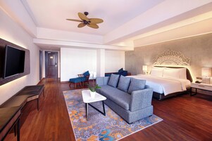 Una camera d'albergo moderna con divano, televisione a schermo piatto, ventola a soffitto, un letto con testiera decorativa e un tavolino con una pianta.