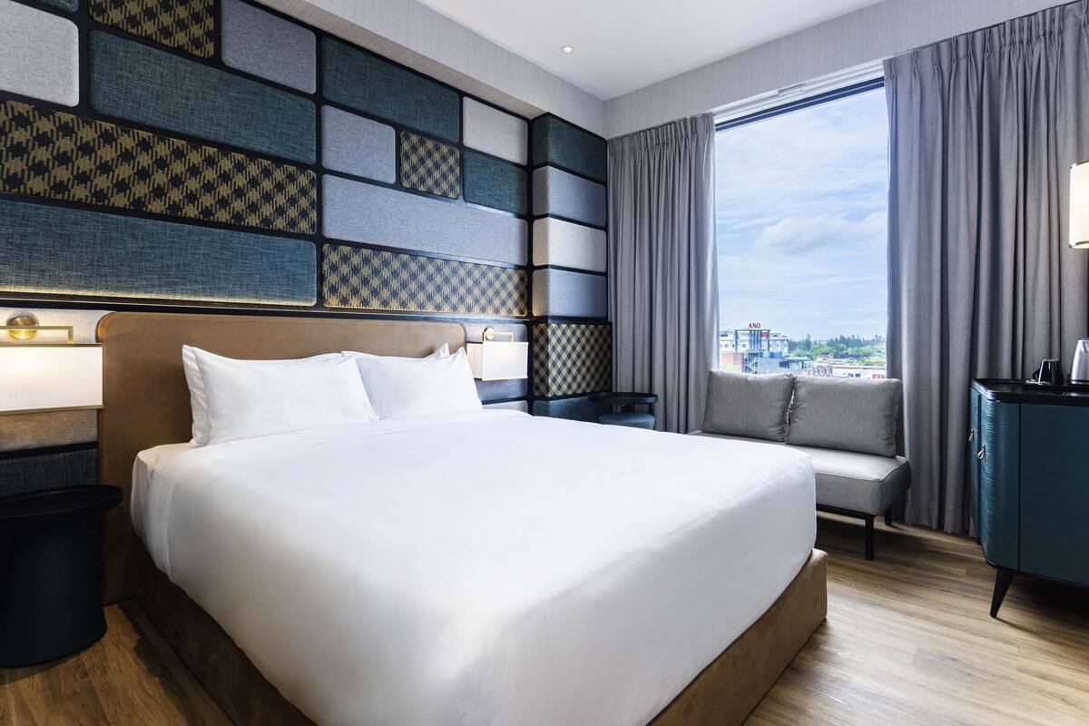 A modern hotel room with a large bed, a seating area, and a view of the city.