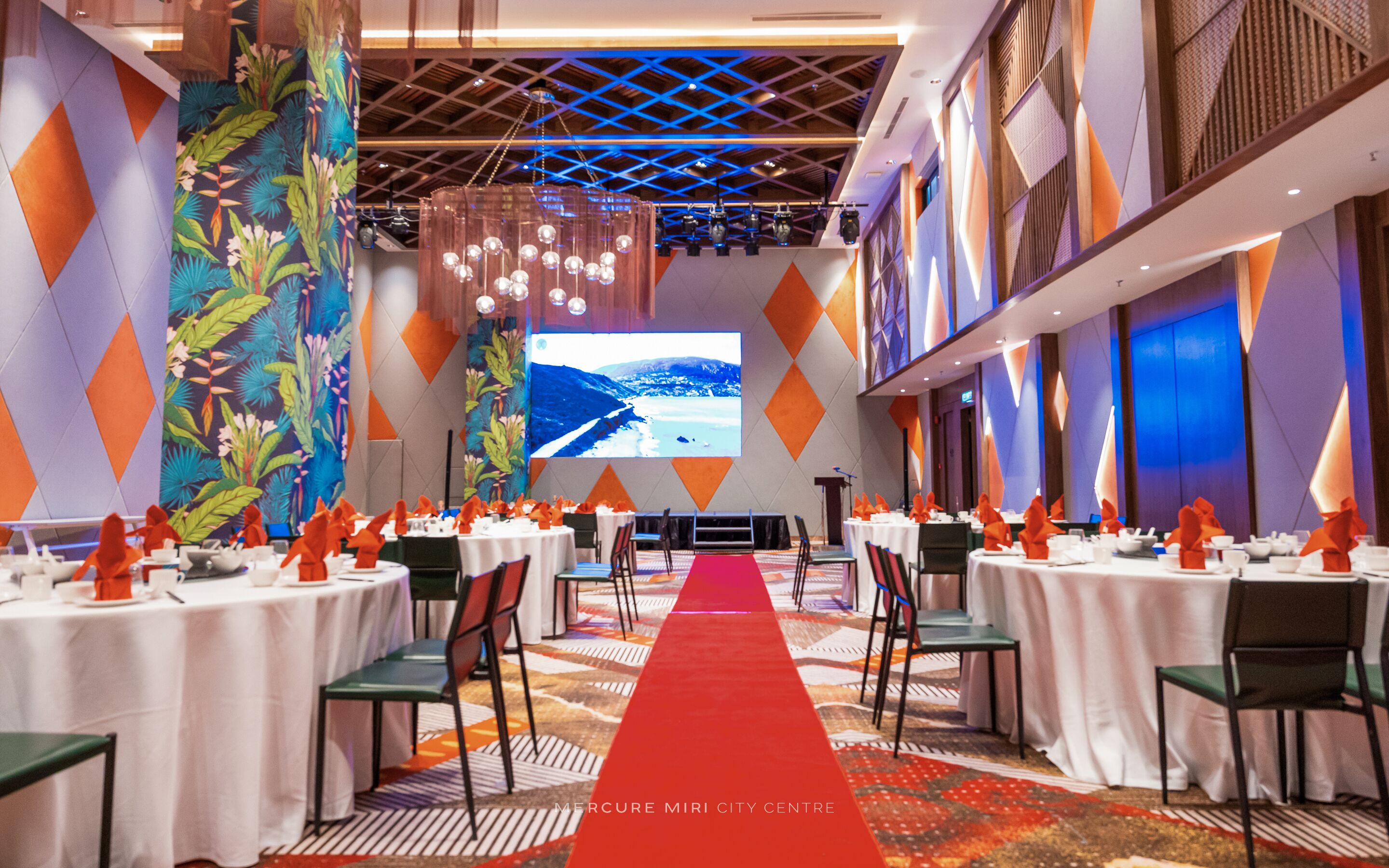 A conference room with a red carpet leading to a stage, tables with white tablecloths, and chairs with orange napkins.