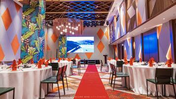 Un salón de conferencias con una alfombra roja que conduce a un escenario, mesas con manteles blancos y sillas con servilletas anaranjadas.