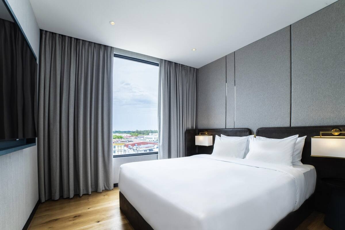 A modern hotel room with a large bed, a flat-screen TV mounted on the wall, and a view of the cityscape through the window.