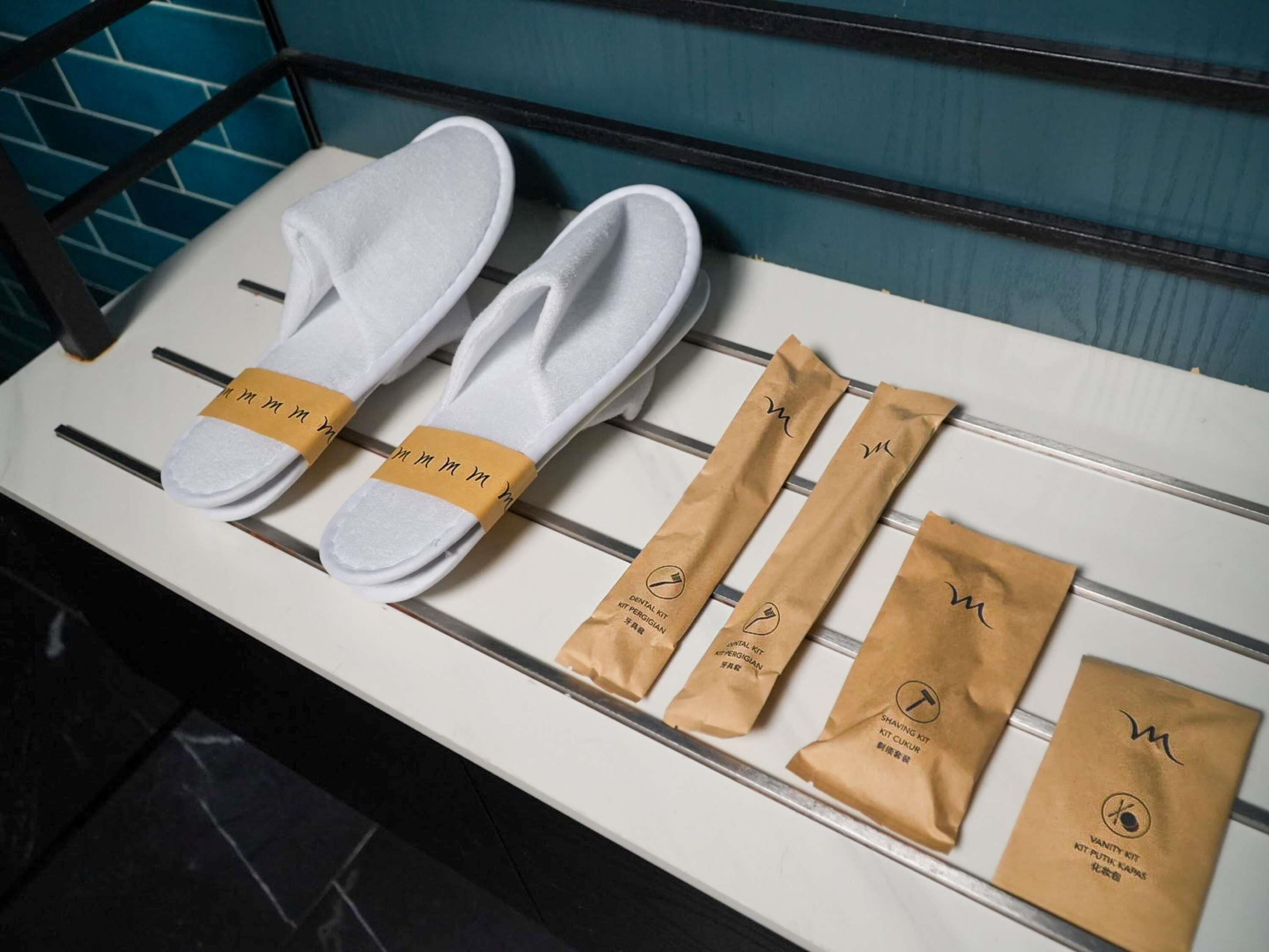 Pantalla mostrando pantuflas blancas y bolsas marrones con texto, dispuestas sobre un estante blanco frente a una pared verde azulado.