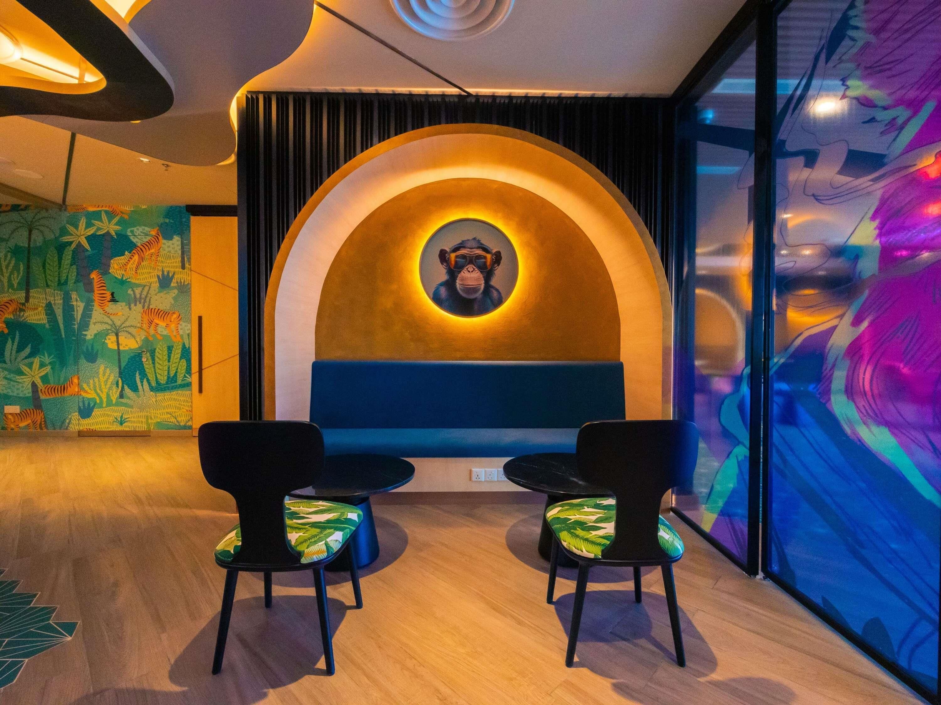 Un interior moderno con una zona de estar que incluye dos sillas negras con cojines de hojas verdes, un banco azul y una pared con un retrato circular de un mono.