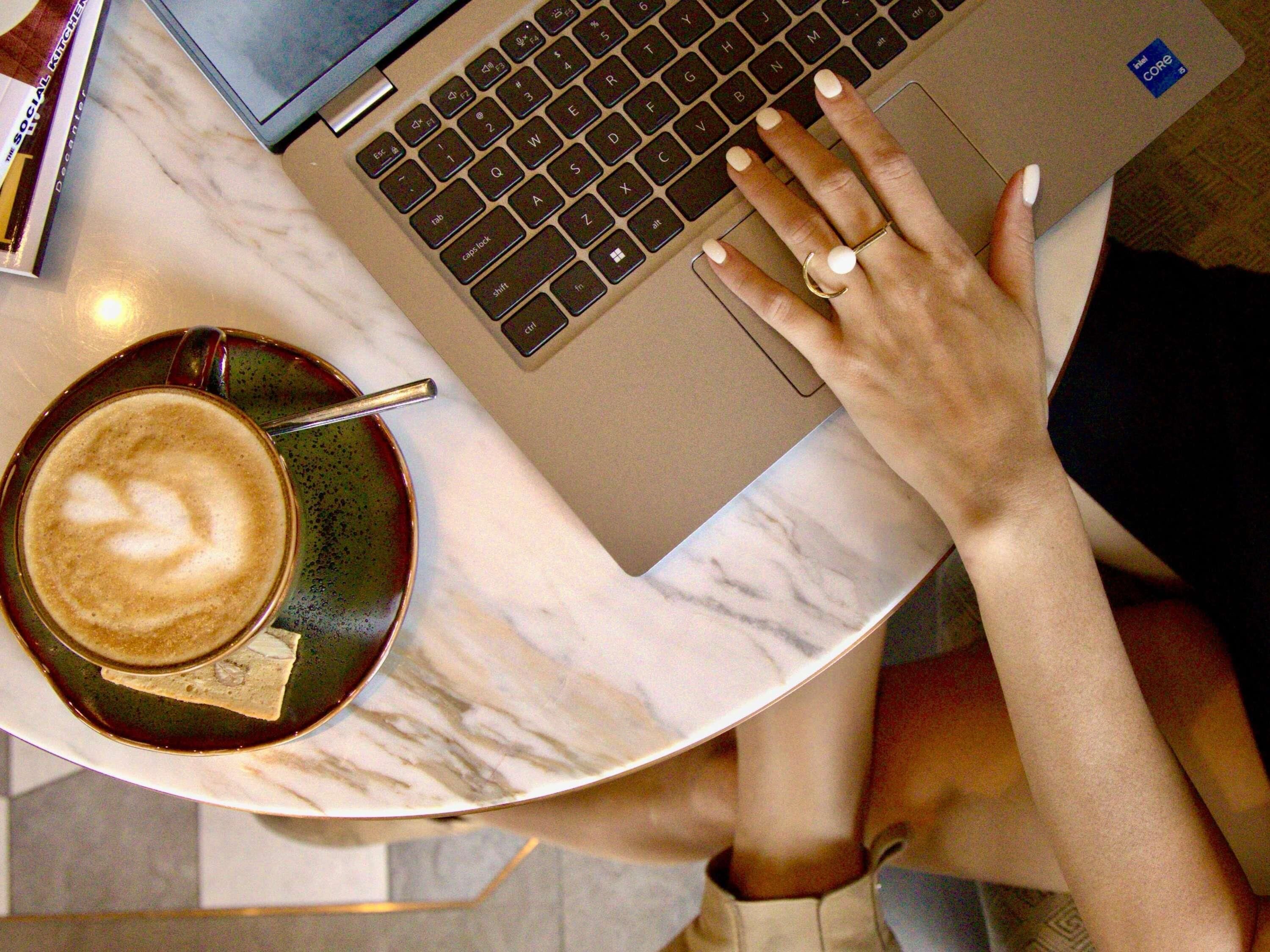 A laptop on a marble surface with a hand resting on it, a cup of coffee with latte art, and a book nearby.