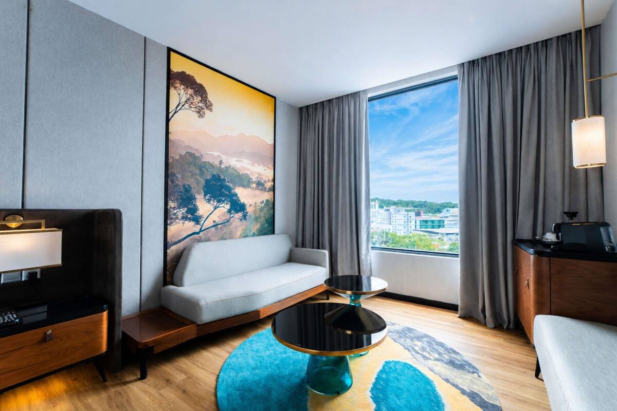 A modern hotel room with a sofa, a small round table, a desk, a lamp, and a large window with curtains.