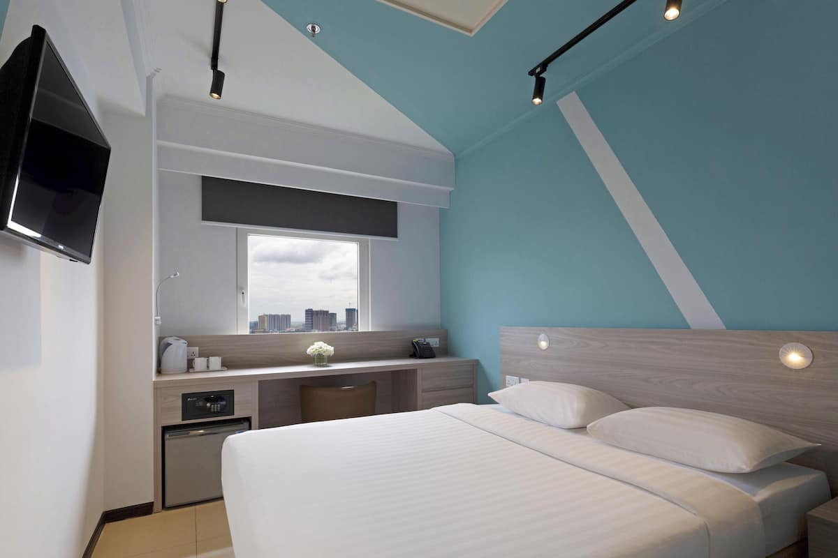 A modern hotel room with a large bed, a desk with a computer, a TV mounted on the wall, and a window with a city view.