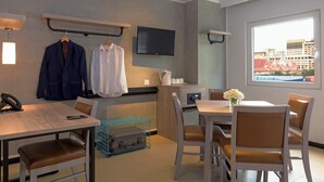 A hotel room with a dining area, a TV mounted on the wall, a window with a city view, and a desk with a phone and a suitcase. - Ibis Budget Phnom Penh Riverside (Phnom Penh)