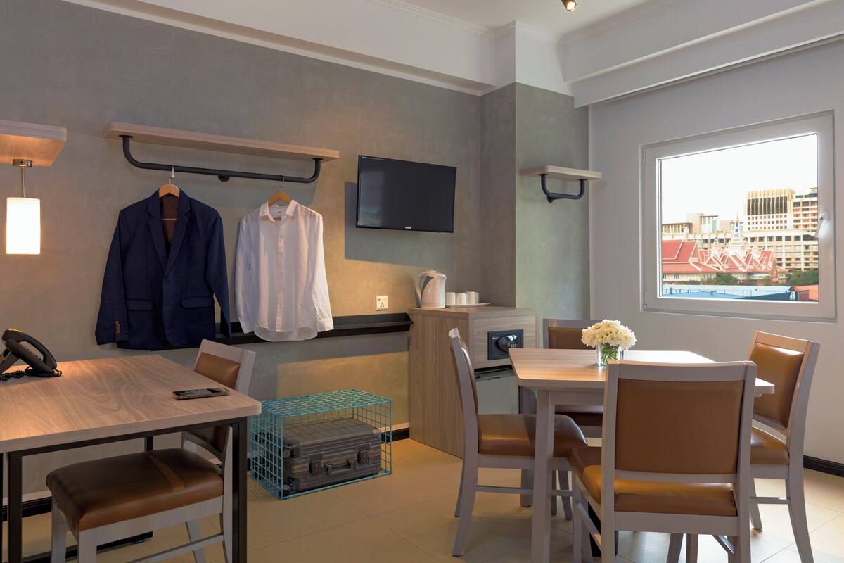 A hotel room with a dining area, a TV mounted on the wall, a window with a city view, and a desk with a phone and a suitcase.